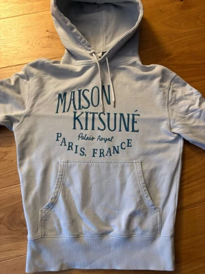Ljusgrå hoodie Maison Kitsuné XS - Snygg ljusgrå hoodie från Maison Kitsuné med tryck i blått framtill där det står 'Maison Kitsuné Paris, France'. Bara använd ett fåtal gånger köpt på miinto för ca 1 år sedan har kvitto också.