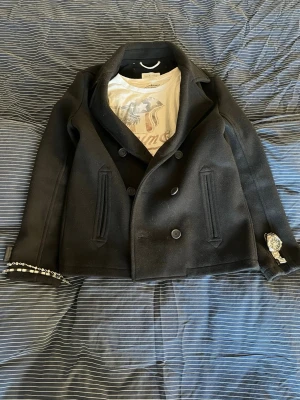 Svart skepparkavaj från Pier One - Klassisk svart skepparkavaj från Pier One med dubbelknäppning och bred krage. Jackan har två snedställda fickor framtill och dekorativa knappar. Materialet känns tjockt och värmande, perfekt för kyligare dagar.