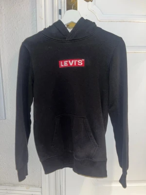 Svart hoodie från Levi's med logga - Svart hoodie från Levi's med klassisk röd och vit logga på bröstet. Hoodien har huva, magficka och ribbade muddar vid ärmslut och nederkant. Tillverkad i mjukt bomullsmaterial, perfekt för en avslappnad och stilren look.
