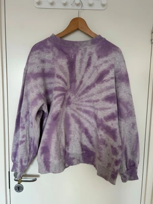 Lila tie-dye hoodie - Säljer en oversized hoodie med lila och grått tie-dye mönster. Tröjan har en avslappnad passform, ribbade muddar och är tillverkad i mjukt bomullsmaterial. Perfekt för afterski