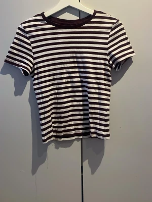 Randig t-shirt från H&M Divided - Säljer en kortärmad t-shirt från H&M Divided med horisontella vinröda och vita ränder. T-shirten har en klassisk rund halsringning och är gjord i mjuk bomull. Perfekt till jeans eller kjol för en chill look.
