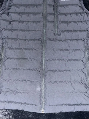 Svart dunväst från Peak Performance - Snygg svart dunväst från Peak Performance med quiltad design och dragkedja framtill. Västen har en bröstficka med dragkedja och två sidofickor. Perfekt för lager-på-lager och håller dig varm med lätt dunfyllning. Perfekt inför våren