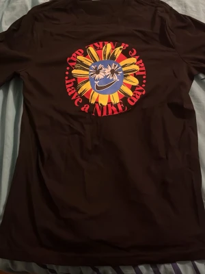 Svart Nike t-shirt med solrosmotiv - Cool svart t-shirt från Nike med stort färgglatt solrosmotiv och texten 'Have a Nike Day' på bröstet. Trycket har gula, röda och blå detaljer samt ett smileyface med Nike-logga. Tillverkad i mjuk bomull och har klassisk passform med korta ärmar.