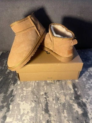 Klassiska beige UGG boots - Säljer ett par klassiska beige UGG boots i mocka med mjukt vitt foder. Skorna har rund tå, låg skaft och grov sula för extra komfort. Perfekta för kalla dagar och riktigt sköna att ha på sig. UGG-logga bak på hälen.