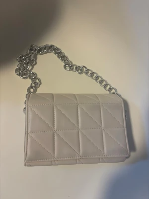 Beige väska med kedja - Snygg beige quiltad handväska med silverfärgad kedjerem. Väskan har ett rutmönster i sömmarna och en rektangulär form. Perfekt accessoar för att lyfta din outfit.