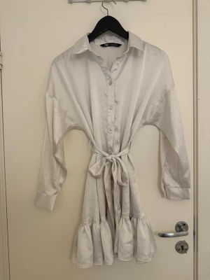 Vit silke klänning med knytband - Stilren vit silke klänning från Zara i glansigt material med knappar framtill, lång ärm och volang nedtill. Knytband i midjan för markerad siluett och klassisk krage. Perfekt för en trendig och avslappnad look.