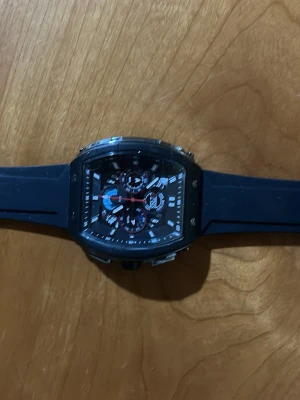 Svart sportig klocka med gummiband - Snygg svart klocka med fyrkantigt ur och flera små urtavlor. Klockan har ett svart gummiband och detaljer i silver och rött på urtavlan. Perfekt accessoar för en modern och sportig stil.