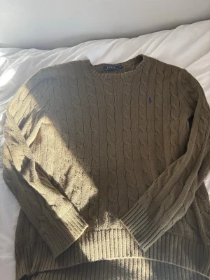 Olivgrön kabelstickad tröja Polo Ralph Lauren - Säljer en olivgrön kabelstickad tröja från Polo Ralph Lauren med rund halsringning och lila broderad logga på bröstet. Tröjan har långa ärmar och ribbade muddar vid ärmslut och nederkant. Perfekt för dig som gillar klassisk och stilren look.pris kan diskuteras vid snabb äffär!