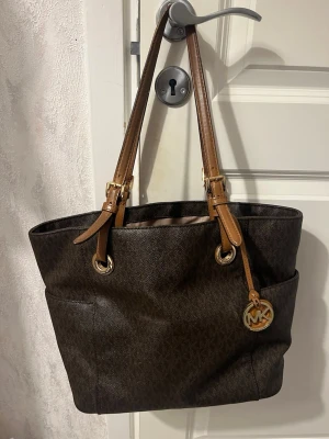 Brun Michael Kors handväska - Snygg brun handväska från Michael Kors med klassiskt MK-mönster och guldfärgade detaljer. Väskan har två långa axelband i ljusbrunt läder och en rund MK-logga som hänge. Praktiska ytterfickor på sidorna och rymlig insida. Perfekt för dig som gillar stilrena accessoarer.