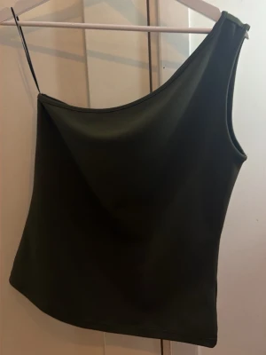 Mörkgrön one shoulder topp - Stilren mörkgrön topp med one shoulder-design. Toppens enkla snitt och släta material ger en modern och clean look. Perfekt att matcha med både jeans och kjol för en trendig outfit.