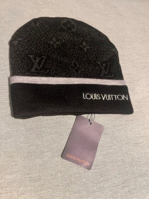Svart grå mössa från L - Snygg svart mössa från L@uis V@itton med klassiskt monogrammönster och grå logotyp framtill. Mössan har en grå rand och är stickad i mjukt material som känns lyxigt. Perfekt accessoar för dig som gillar exklusiv streetstyle.