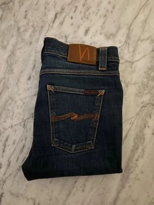 Nudie jeans - Snygga mörkblå skinny jeans från Nudie med klassiska orange sömmar och läderpatch bak i midjan. Jeansen har fem fickor och en smal passform som sitter tajt längs benen. Perfekta för en trendig och avslappnad stil.