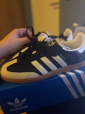 Adidas Samba OG svart/vit sneaker - Klassiska Adidas Samba OG sneakers i svart och vitt med de ikoniska tre ränderna på sidan. Ovandel i mocka och läder, gummisula och svart snörning. Tidlös och sportig design som funkar till det mesta. Modellnamnet 'SAMBA' syns i silver på sidan.