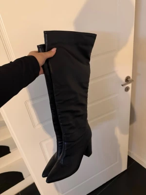 Svarta knähöga boots med klack - Säljer ett par svarta knähöga boots med spetsig tå och bred klack. Stövlarna har en slät finish och är tillverkade i ett material som liknar skinn. Perfekta för att lyfta vilken outfit som helst.