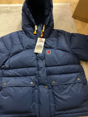 Blå dunjacka med huva från Fjällräven - Snygg blå dunjacka från Fjällräven med stor huva, gula dragskor och två stora fickor med lock och tryckknappar. Jackan har Fjällrävens klassiska logga på bröstet och är perfekt för kalla vinterdagar. Materialet är slitstarkt och isolerande.