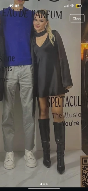 Svart satinklänning med scarfdetalj Zara  - Stilren svart klänning i satin med lång ärm och lös passform. Djup v-ringning och dekorativ scarfdetalj vid halsen. Klänningen har en lätt glansig yta och slutar ovanför knäna. Perfekt för dig som gillar en trendig och elegant look.