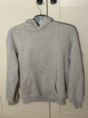 Grå basic hoodie med huva - En enkel och stilren grå hoodie med huva och magficka. Tillverkad i mjukt sweatshirtmaterial som är skönt att ha på sig. Perfekt för en avslappnad look och funkar till det mesta. Klassisk passform och ribbade muddar vid ärmar och nederkant.