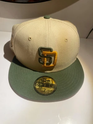 San Diego Padres 59FIFTY keps beige/grön, Size 7 - Säljer en San Diego Padres 59FIFTY keps från New Era i beige med grön skärm. Framtill finns SD-loggan i grönt och gult, och på sidan ett broderat 1992 All Star Game-märke. Klassisk flat brim och broderade ventilationshål. Perfekt för dig som gillar sportig stil.