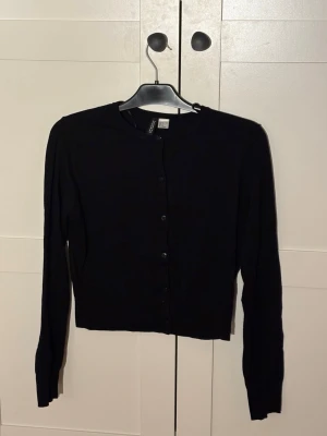 Svart cardigan från H&M - Klassisk svart cardigan från H&M med rund halsringning och knäppning framtill. Långärmad och i mjukt, tunt material som passar perfekt till lager på lager. Enkel och stilren design som funkar till det mesta.
