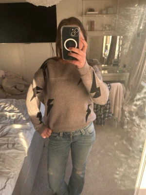 Beige stickad tröja med blixtmotiv - Cool beige stickad tröja med svarta blixtar som detaljer. Tröjan har rund halsringning och långa ärmar. Perfekt för dig som vill ha en unik och trendig look till vardags. Materialet är mjukt och bekvämt. 