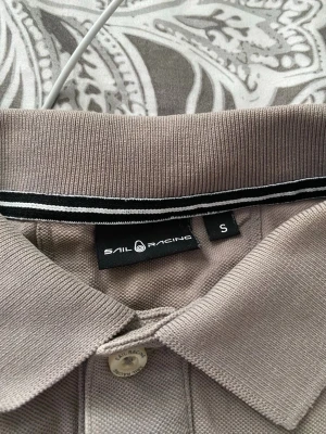 Beigegrå pikétröja från Sail Racing - Säljer en beigegrå pikétröja från Sail Racing med klassisk krage och knappslå framtill. Tröjan har broderad logga på bröstet och svarta/vita detaljer på kragen. Perfekt för en clean och sportig stil.