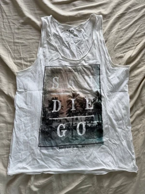 Vit linnetopp med San Diego-tryck från Smog - Säljer en vit ärmlös linnetopp från Smog med ett stort San Diego-tryck framtill med palmmotiv. Toppen har avslappnad passform och rund halsringning. Perfekt för varma dagar eller som ett chill plagg till stranden.