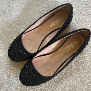 Ballerinaskor - Svarta ballerinaskor med glittriga stenar över hela ovandelen. Skorna har rund tå och platt sula, vilket gör dem både snygga och bekväma. Perfekta för dig som vill ha lite extra bling i vardagen.