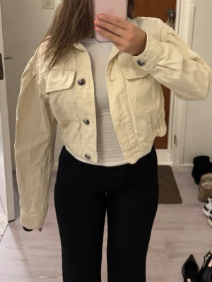 Ljusbeige cropped jeansjacka - Trendig ljusbeige cropped jeansjacka med stora bröstfickor och silverfärgade knappar. Jackan har en boxy passform och elastisk midja för en snygg siluett. Perfekt att slänga över en t-shirt för en cool och avslappnad look.