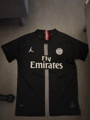 PSG Neymar Jr matchtröja svart - Snygg svart PSG matchtröja med Neymar Jr och nummer 10 på ryggen. Tröjan har Fly Emirates-tryck på bröstet, PSG-logga och Jordan-logga. Röd och vit rand på kragen och en vertikal grå rand framtill. Perfekt för fotbollsfans!