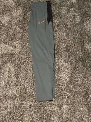 Grå Nike mjukisbyxor med orange logo - Snygga grå mjukisbyxor från Nike med en orange swoosh-logga på låret och svarta detaljer vid fickan. Byxorna har en smal passform och resår i midjan. Perfekta för chill eller träning.