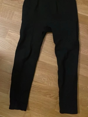 Svarta träningsbyxor  - Ett par svarta träningsbyxor med hög midja och smal passform. De är stretchiga och mjuka, perfekta för en avslappnad stil. Materialet ser ut att vara bomull eller en bomullsblandning. Inga synliga detaljer eller loggor.