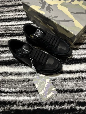 Svarta sneakers från Valentino Garavani - Snygga svarta sneakers från Valentino Garavani med diskreta kamouflagedetaljer och svart sula. Skorna har en klassisk låg profil, svarta skosnören och är tillverkade i en mix av läder och textil. Perfekta för dig som gillar stilrena och exklusiva sneakers.