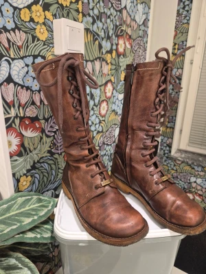 ANGULUS läder, platta, med snören, Långa stövlar Brun Storlek 38 - Material: 100% läder Yttersula: fsc-certifierat svart crepegummi så Tillverkad i Portugal  Produktinformation Spetsdesignen möter en platt sula i denna mångsidiga känga. Den höga axeln ger utökad täckning, medan den främre spetsstängningen säkerställer en säker och justerbar passform.  Viktiga funktioner Justerbar snörning Platt sula Egenskaper Högt skaft Spetsen framifrån  Dessa stövlar är perfekta för kvinnor som vill ha ett snyggt och bekvämt alternativ för vardagsbruk.