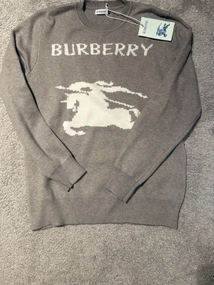 Grå stickad tröja från Burberry - Snygg grå stickad tröja från Burberry med vit logga och ikonisk riddare framtill. Tröjan har rund halsringning, långa ärmar och ribbade muddar. Perfekt för dig som vill ha en klassisk men ändå trendig look.