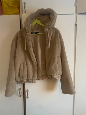 Beige teddyjacka med huva från Zara - Mysig beige teddyjacka från Zara med stor huva och dragsko. Jackan har två fickor framtill, ribbad nederkant och är fodrad på insidan för extra komfort. Perfekt för kyliga dagar och ger en trendig look.