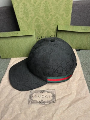 Svart Gucci keps med GG-mönster - Snygg svart keps från Gucci med det klassiska GG-monogrammet över hela tyget. Kepsen har en böjd skärm och en ikonisk grön-röd rand på sidan. Tillverkad i canvasmaterial som ger en exklusiv känsla och streetwear-vibe.