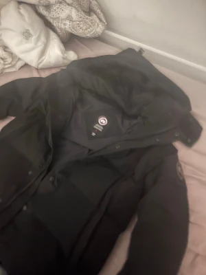 Svart dunjacka från Canada Goose - Säljer en svart dunjacka från Canada Goose med huva och klassisk logotyp på ärmen. Jackan har dragkedja och knappar och är i storlek M fast passar även S