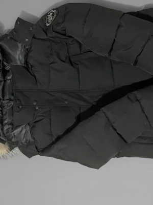 Svart lång pufferjacka med fuskpäls - Svart pufferjacka med lång passform och huva med fuskpäls. Jackan har quiltad design, tryckknappar och dragkedja framtill samt ett emblem på axeln. Fodret är glansigt och mjukt för extra komfort. Perfekt för kalla dagar. Pris kan diskuteras vid snabb affär.