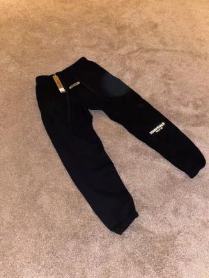 Svarta Essentials mjukisbyxor - Säljer ett par svarta mjukisbyxor från Fear of God Essentials med logga tryckt på benet. Byxorna har resår i midjan och vid bensluten samt dragsko för bästa passform. Perfekta för en chill och trendig look.