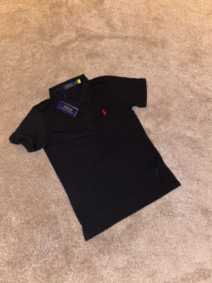 Svart pikétröja från Polo Ralph Lauren - Klassisk svart pikétröja från Polo Ralph Lauren med röd broderad logga på bröstet. Tröjan har korta ärmar, krage och knappar framtill. Tillverkad i mjuk bomull som känns skön mot huden. Perfekt för en stilren och avslappnad look.