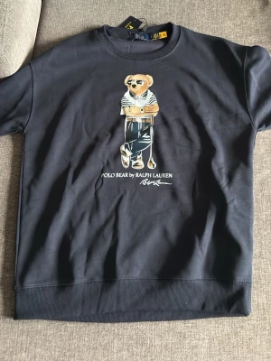 Svart sweatshirt Polo Ralph Lauren - Cool svart sweatshirt från Polo Ralph Lauren med den ikoniska Polo Bear-trycket på bröstet. Tröjan har rund halsringning, ribbade muddar och är gjord i mjukt bomullsmaterial. Perfekt för en avslappnad och trendig look. Denna tröja är helt ny där lapparna hänger kvar. Denna fungerar till dem som har L i storlek och även M.