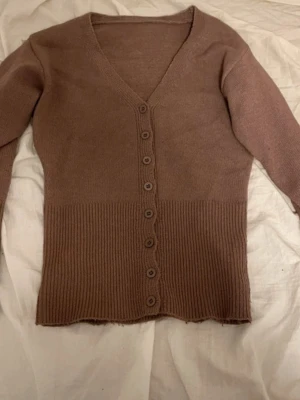 Brun ribbstickad kofta med knappar - Mysig brun kofta med v-ringning och långa ärmar. Koftan har ribbstickad nederdel och knäppning med sju knappar framtill. Perfekt för lager-på-lager och enkel att matcha med olika outfits.