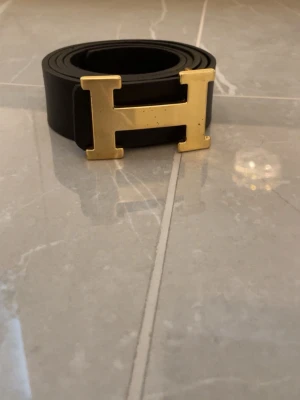 Svart bälte med guldfärgat H-spänne - Snyggt svart bälte  med ikoniskt H-spänne i guldfärgad metall. Bältet är gjort i skinn och har en stilren, klassisk design som passar till det mesta. Perfekt accessoar för att lyfta din outfit.