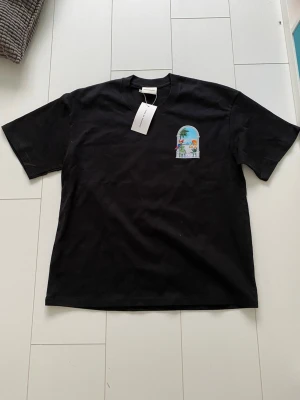 Drole De Monsieur Tshirt med Tryck - Svart t-shirt med färgglatt tryck på bröstet som visar en papegoja, palmer och strandmotiv i ett fönster. Klassisk passform och rund halsringning. Perfekt för dig som gillar stilrena plagg med en twist.