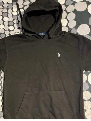 Svart hoodie från Polo Ralph Lauren - Snygg svart hoodie från Polo Ralph Lauren med klassisk vit broderad logga på bröstet. Hoodien har känguruficka framtill och justerbar huva med snören. Tillverkad i mjukt bomullsmaterial som är skönt att bära.
