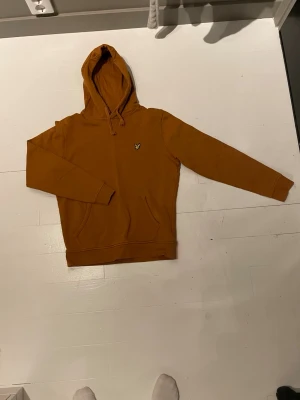 Brun hoodie från Lyle & Scott - Snygg brun hoodie från Lyle & Scott. Har en känguruficka framtill och den ikoniska loggan broderad på bröstet. Tillverkad i mjukt bomullsmaterial som är skönt att bära. Perfekt för en avslappnad och trendig stil. Har dn fläck mot på 