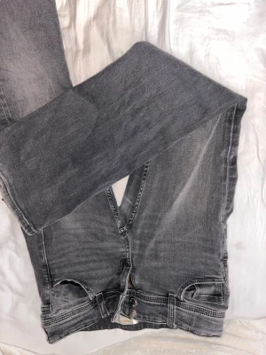 Grå bootcut jeans - Säljer ett par grå bootcut jeans med snygga slitningar och tvättade detaljer. Jeansen har klassisk femficksmodell, dragkedja och knapp i midjan. Perfekta för dig som gillar en avslappnad men trendig look.