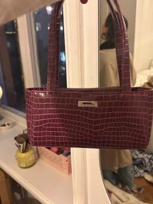 Vintage guess väska - Säljer en lila vintage guess handväska i avlång modell med croco-mönstrad yta och två smala handtag. Väskan har en metallbricka framtill och är tillverkad i glansigt konstläder. Perfekt accessoar för att lyfta din outfit. Köpt i Paris
