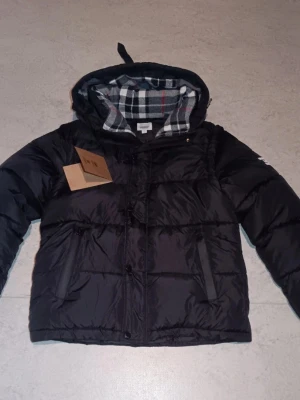 Svart pufferjacka med rutigt foder - Snygg svart pufferjacka med huva och rutigt foder i grått, vitt och rött. Jackan har dragkedja, två sidofickor och en klassisk boxig passform. Perfekt för kalla dagar och riktigt skön att bära. Materialet är glansigt och ger en modern look.