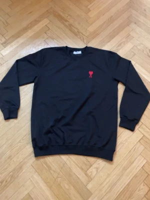 Svart tröja från Ami Paris - Svart sweatshirt från Ami Paris med klassisk rund hals och ribbade muddar. På bröstet finns det ikoniska röda Ami de Coeur-broderiet. Tröjan är tillverkad i mjuk bomull och har en avslappnad passform som passar perfekt till jeans eller byxor. Väldigt billigt pris.